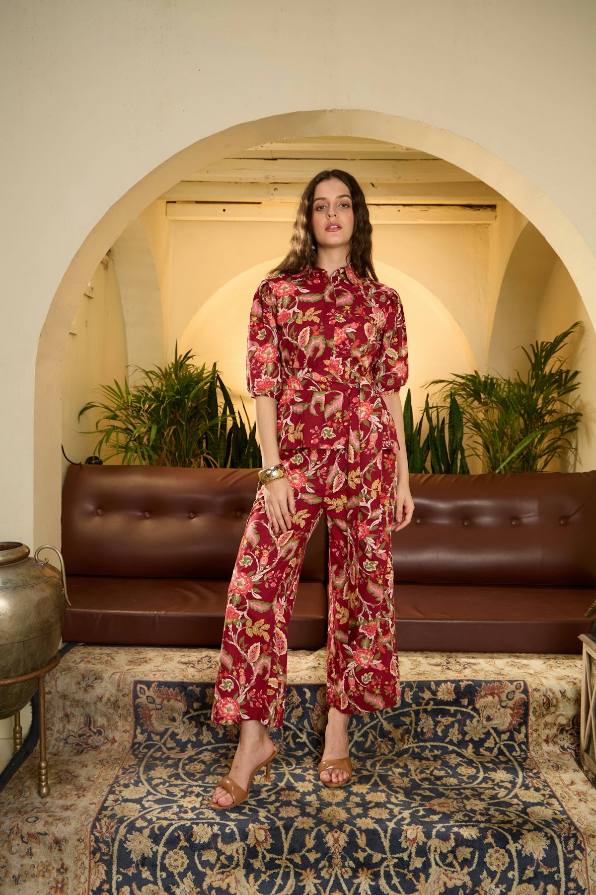Woman in a red floral jumpsuit stands indoors.