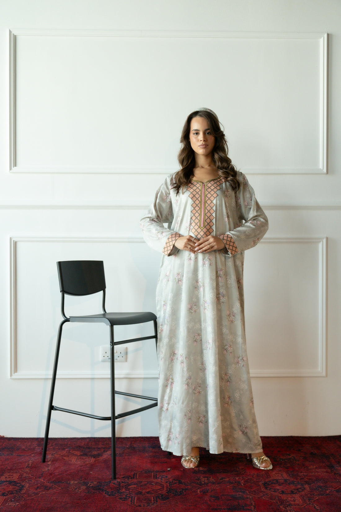 Woman in a long patterned dress stands near a stool.