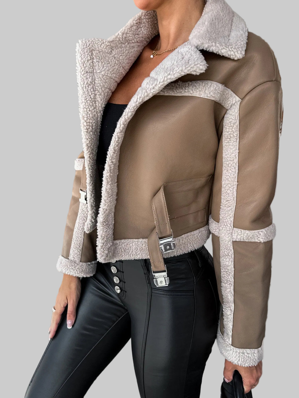 A woman wearing a cropped shearling-lined jacket in taupe and cream, paired with black faux-leather pants, photographed in a concrete indoor setting.