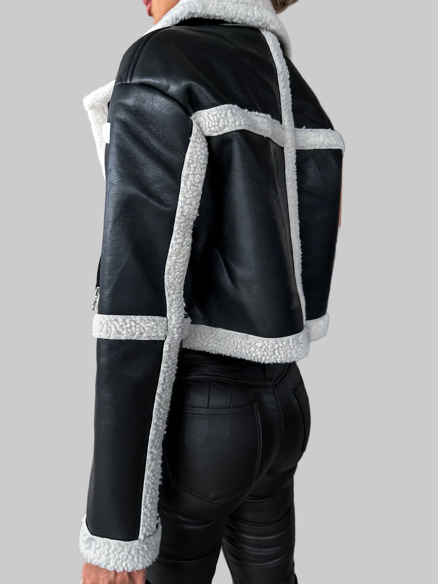 A person shown from the back wearing a black faux-leather jacket with contrasting white sherpa lining on the collar, cuffs, and body panels, paired with black leather pants in an indoor concrete structure.