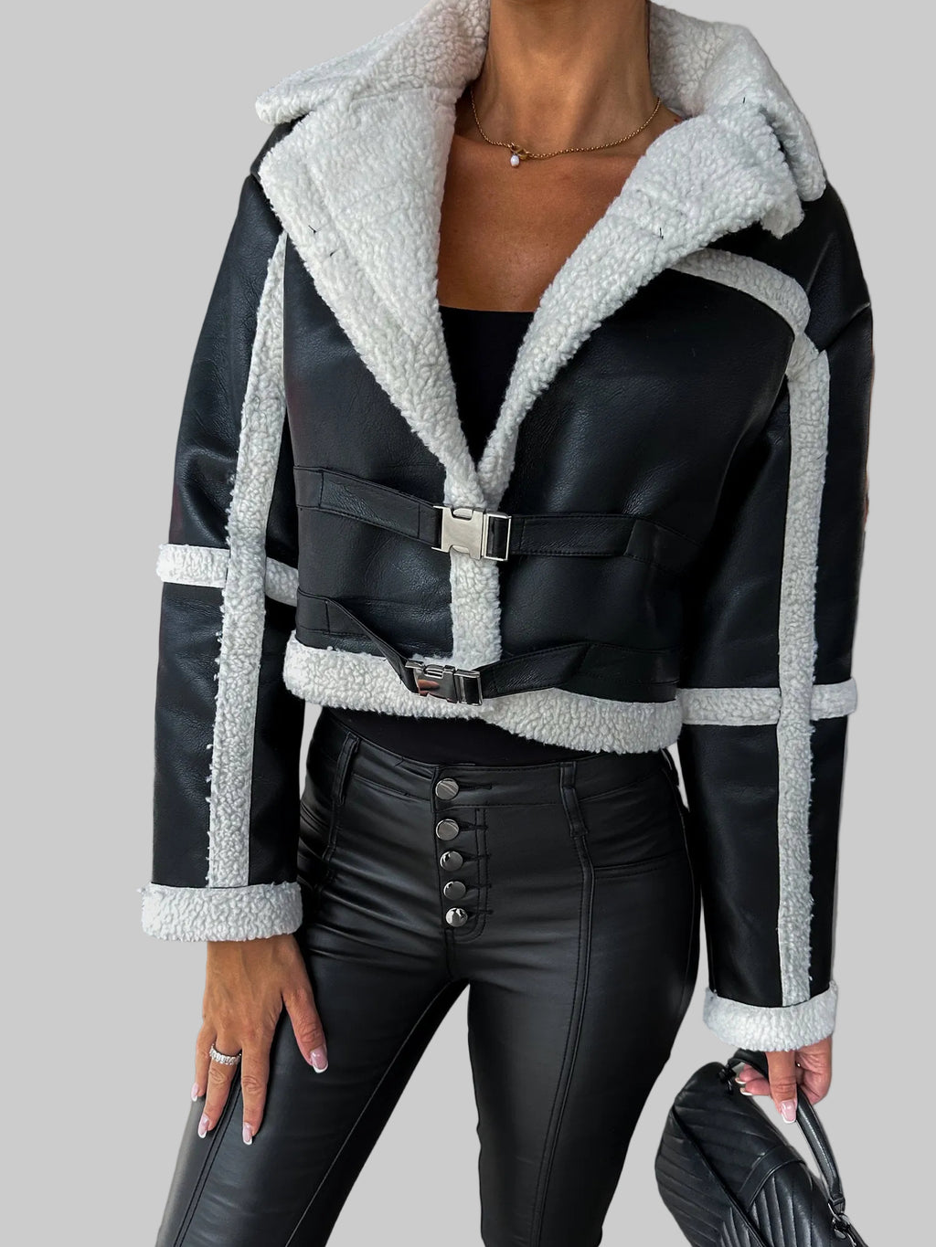 A cropped black faux-leather jacket with a thick white sherpa lining and silver buckle details, worn by a model in a concrete structure.