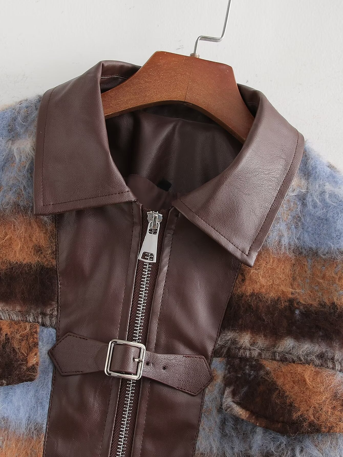 A brown faux-leather jacket with a wide collar, front zipper, and belt buckle detail, hanging on a wooden hanger over a fuzzy blue and brown striped garment.