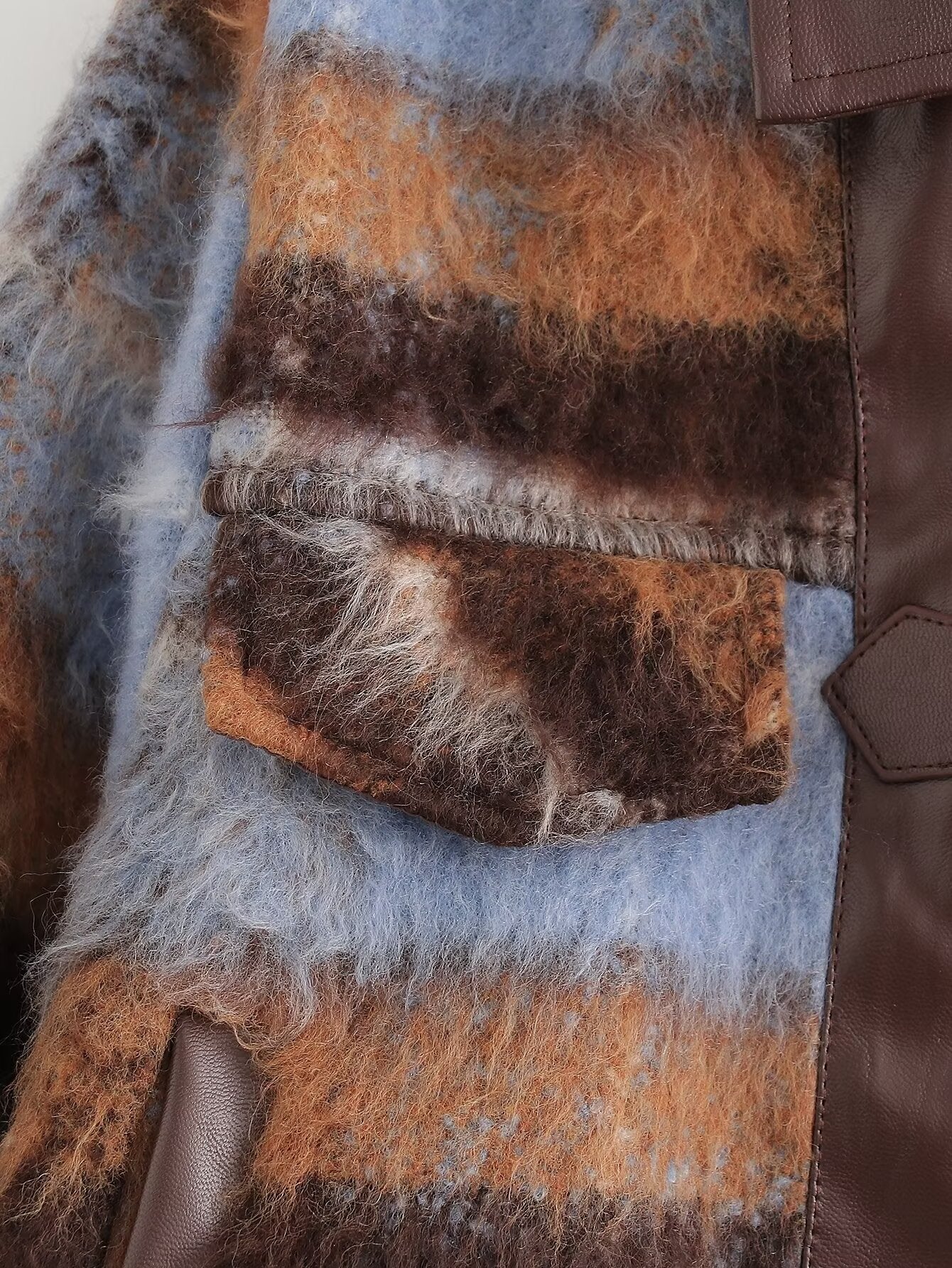 Close-up of a textured patchwork garment featuring fuzzy brown and orange plaid wool-like fabric, light blue wool, and smooth dark brown leather trim.
