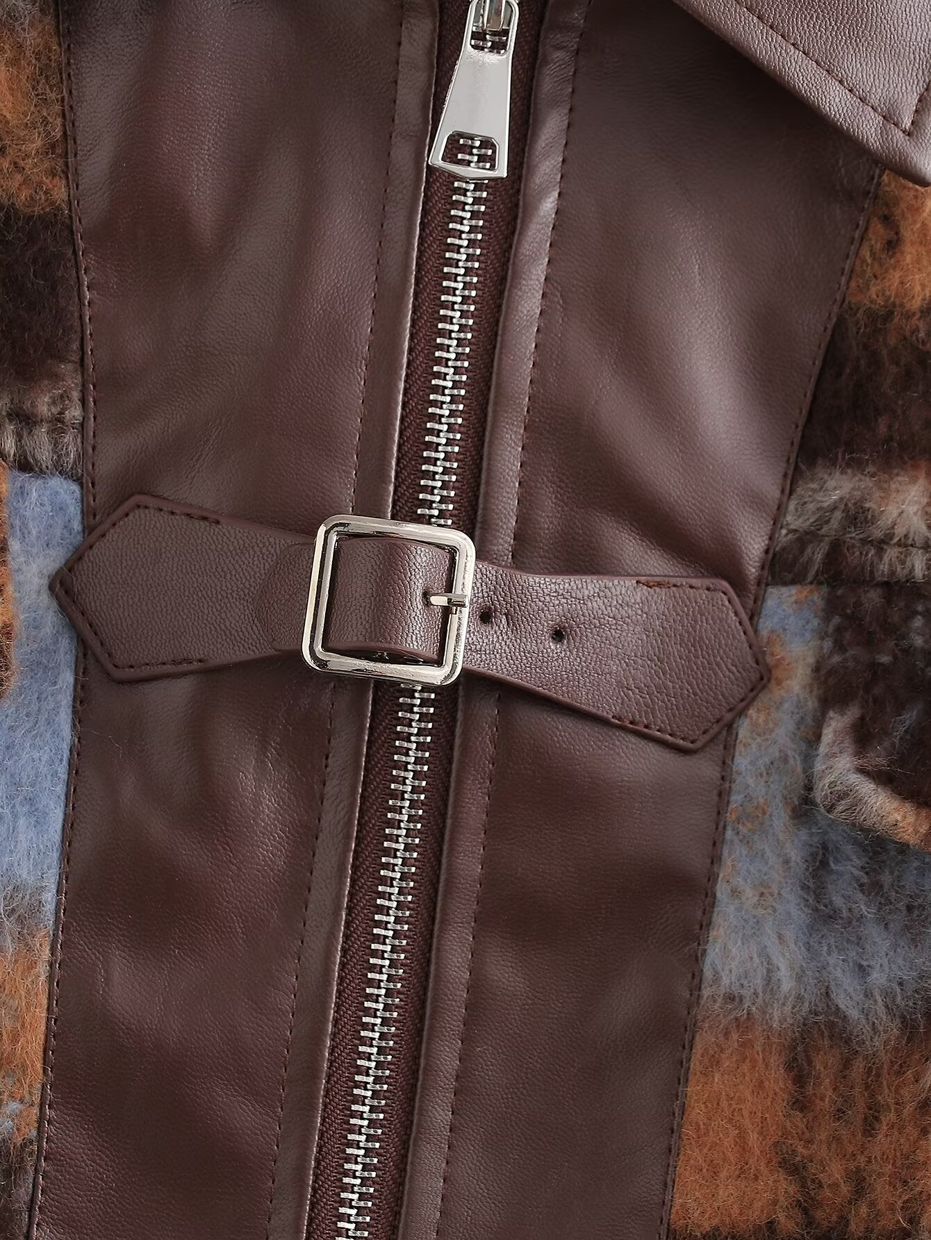 Close-up of a brown faux-leather jacket featuring a central silver zipper, a matching horizontal strap with a square buckle, and a brown and blue patterned faux-fur lining visible at the sides.