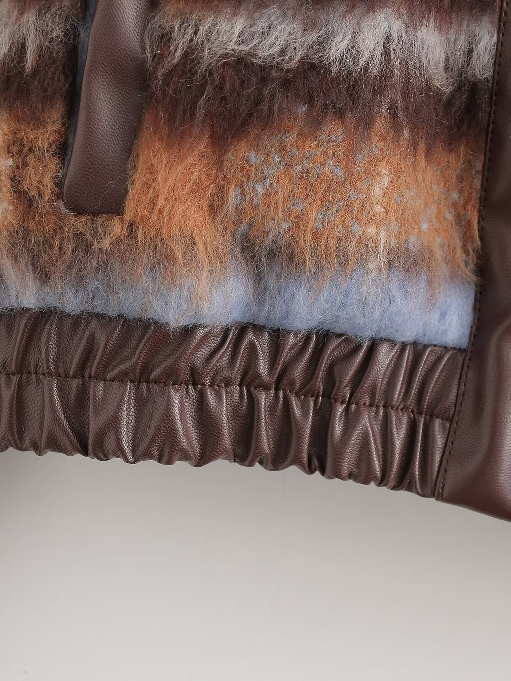 Close-up of a textured handbag featuring a brown faux-leather trim, a gathered elastic bottom, and a fuzzy layered top panel in brown, orange, and light blue-gray.