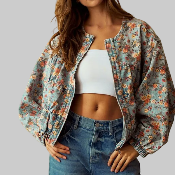 A woman wearing a light blue floral bomber jacket over a white crop top and blue jeans, standing against a warm beige background.