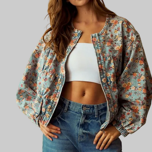 A woman wearing a light blue floral bomber jacket over a white crop top and blue jeans, standing against a warm beige background.