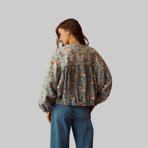 A woman viewed from behind wearing a dusty blue floral cropped jacket with voluminous sleeves and blue jeans, standing in front of a warm beige textured wall.