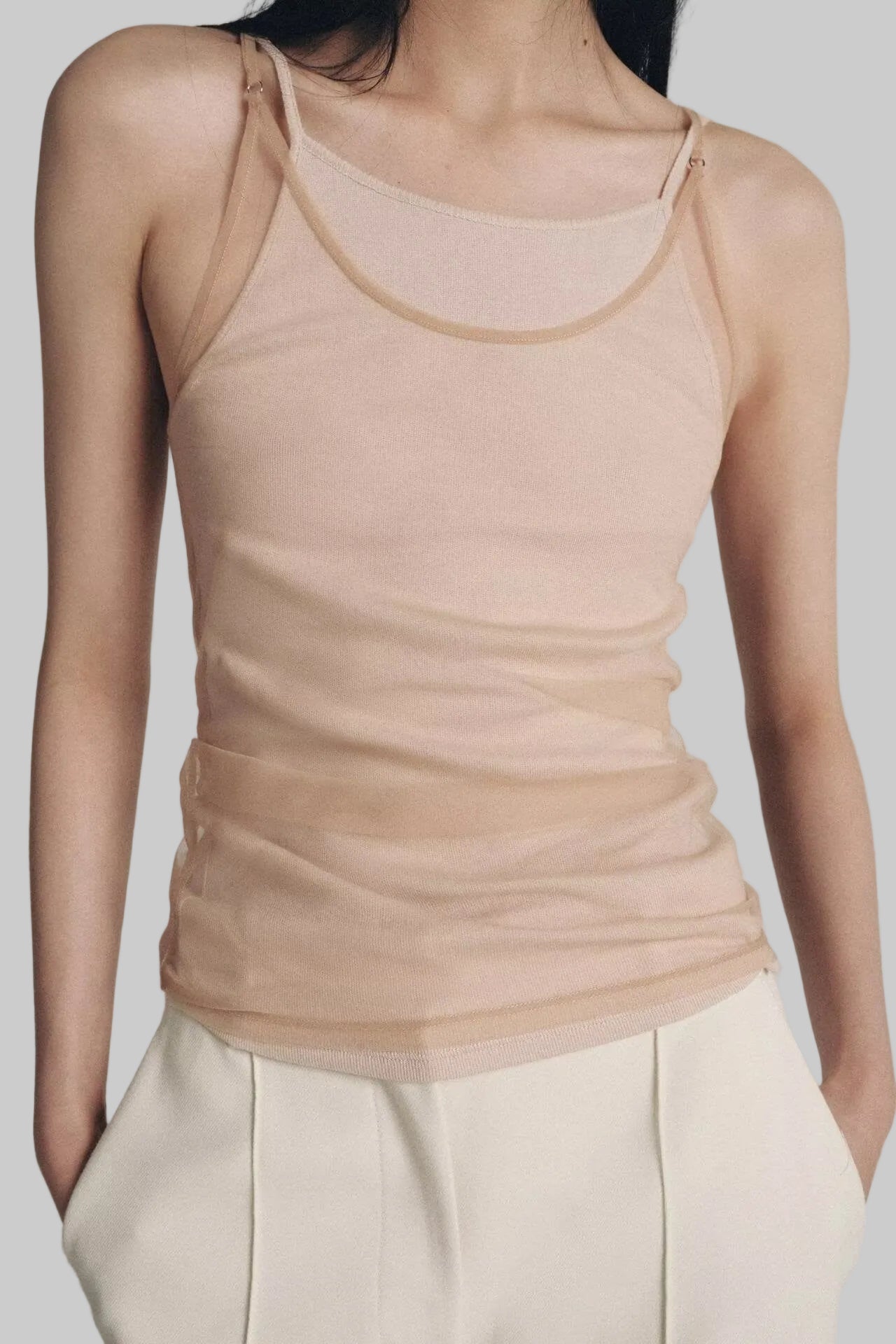 Person wearing a beige tank top and white pants against a neutral background