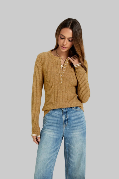 A woman wearing a ribbed knit henley top in camel brown and blue jeans, standing against a light gray studio background.