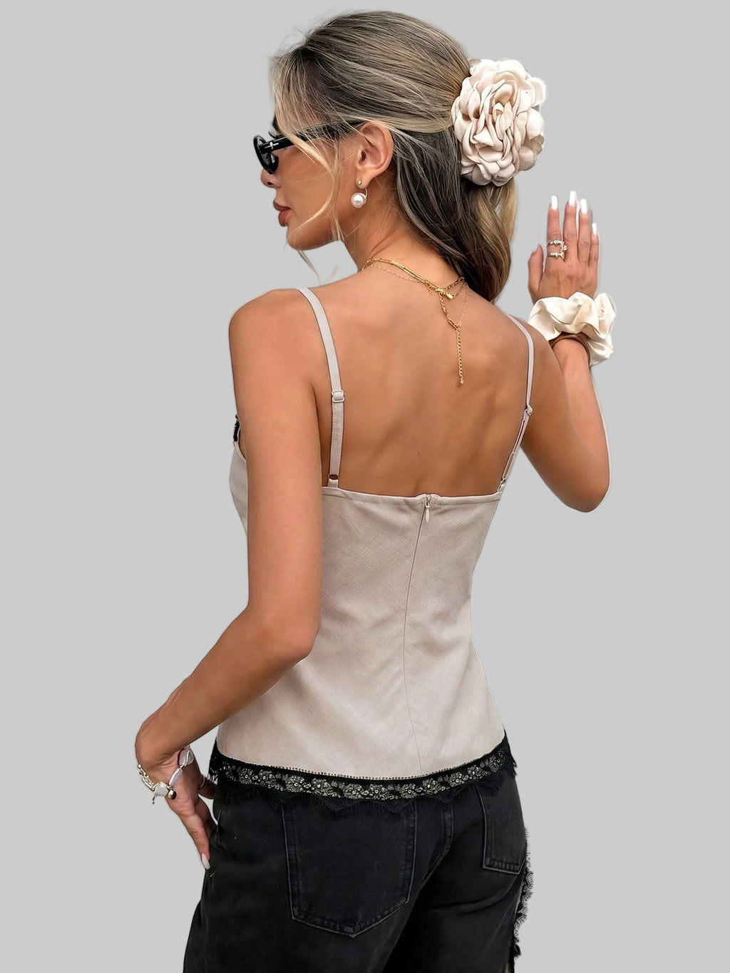 Woman wearing a beige top with thin straps and black pants, standing against a white wall.