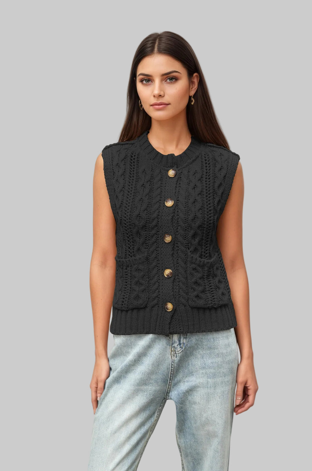 A woman modeling a sleeveless black cable-knit vest with gold-tone buttons, paired with light-wash blue jeans, against a solid light beige background.