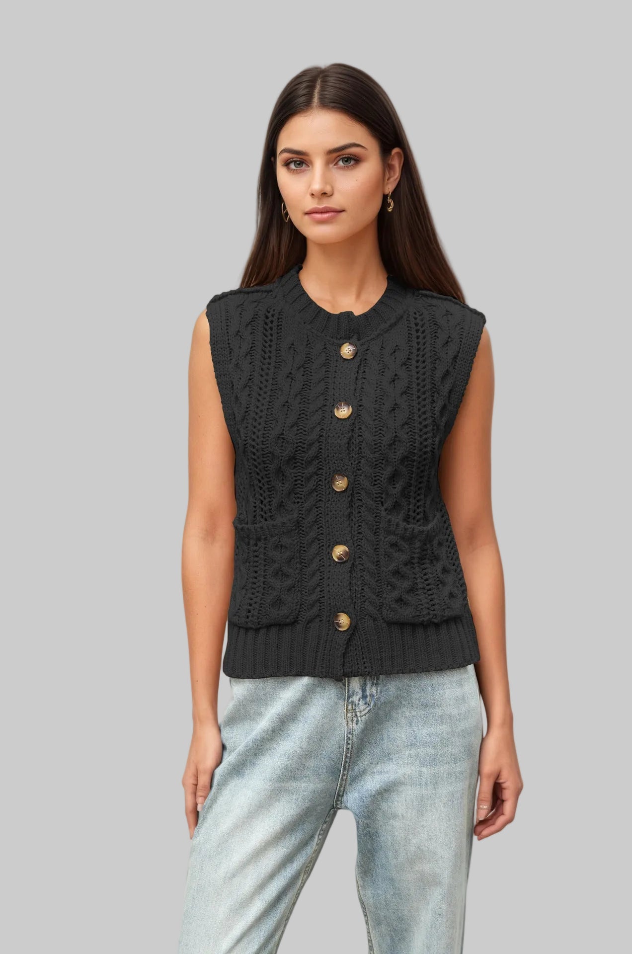 A woman modeling a sleeveless black cable-knit vest with gold-tone buttons, paired with light-wash blue jeans, against a solid light beige background.