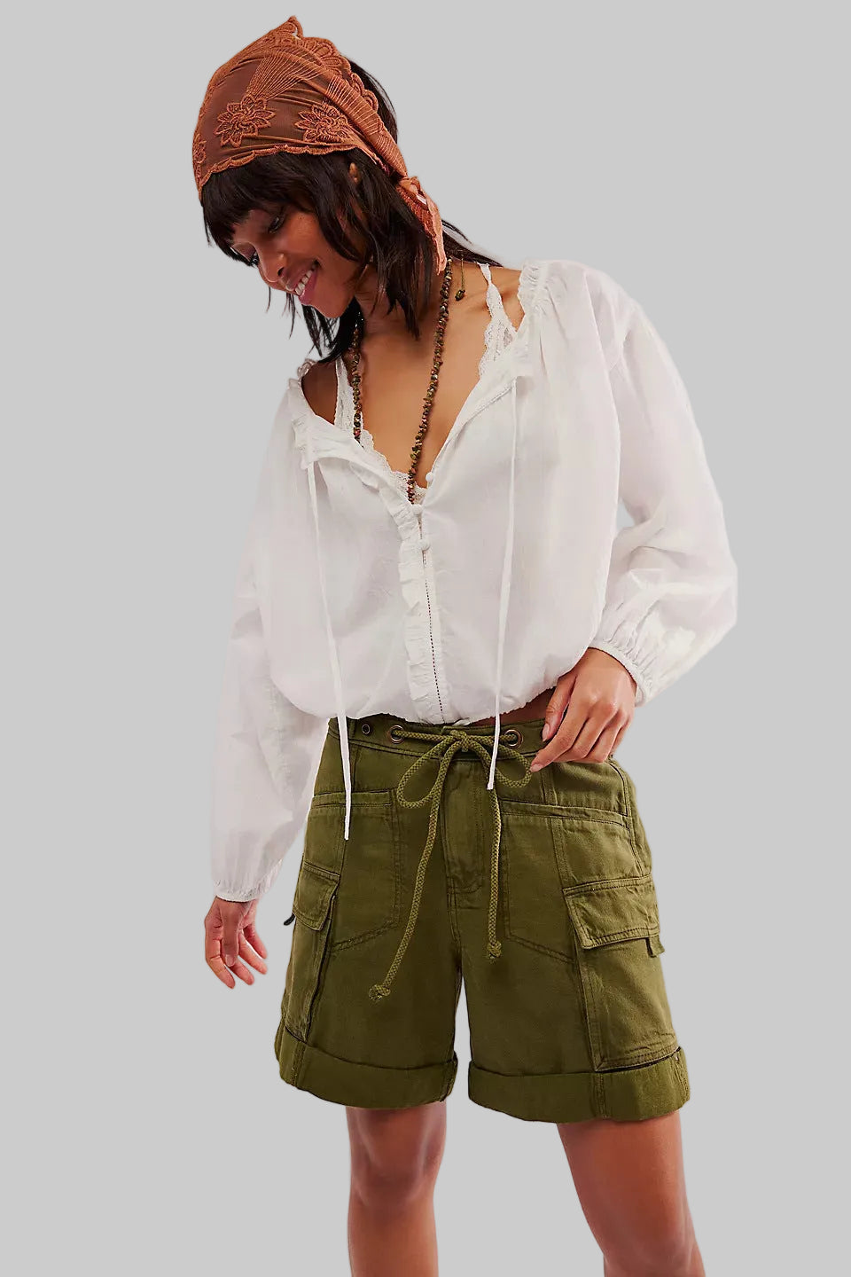 Person wearing a white blouse and green cargo shorts with a brown headscarf on a plain background