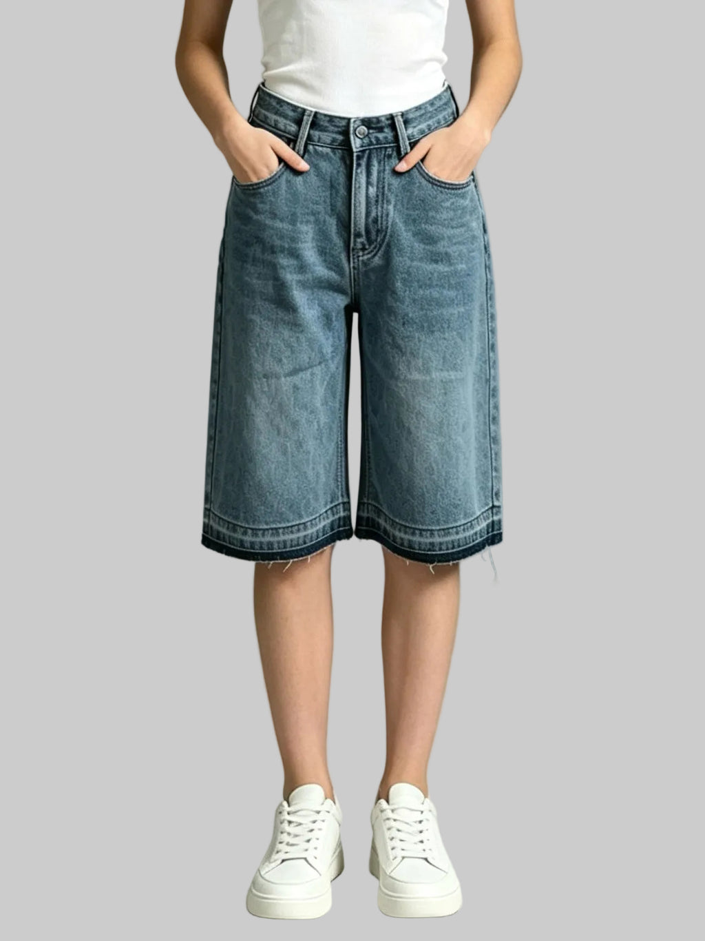 A person wearing light-to-medium wash denim shorts with a frayed hem, paired with a white top and white sneakers, standing against a neutral gray studio background.