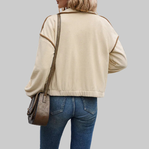 Person wearing a beige jacket and blue jeans with a brown bag, walking on a street.