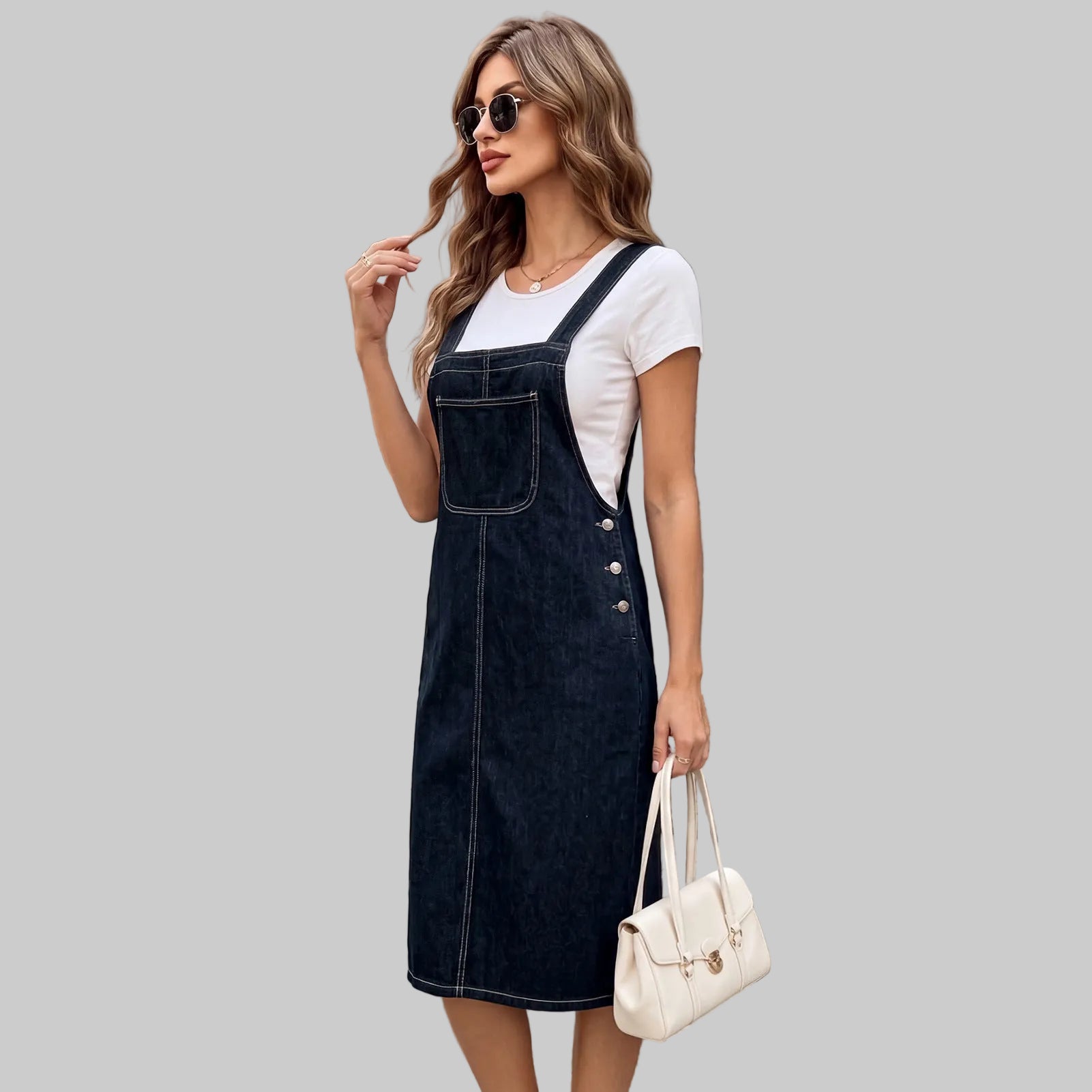 A woman wearing a dark wash denim overall dress over a white t-shirt, standing on stone steps in front of a classic European building. She is holding a white handbag.