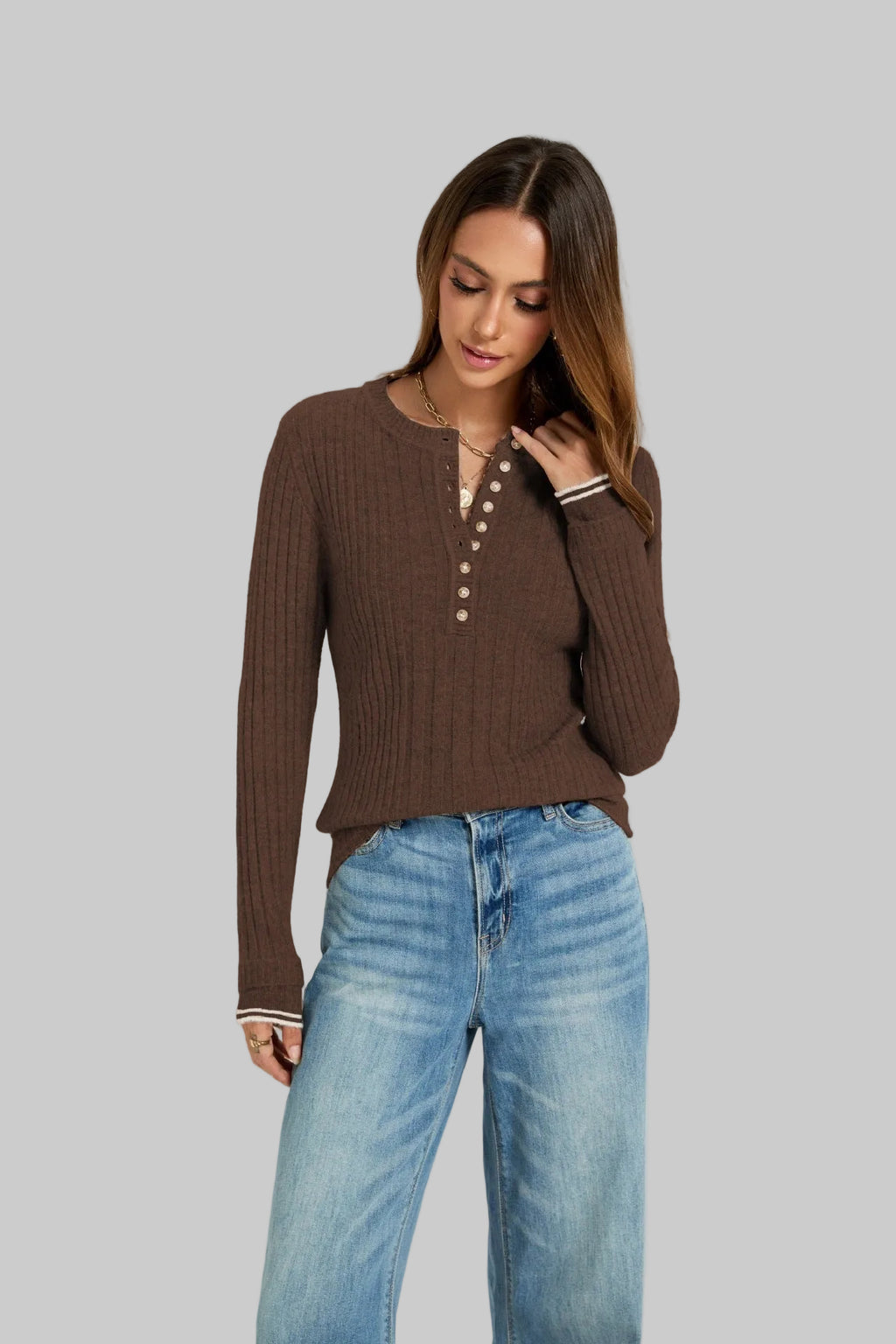 A woman wearing a brown ribbed long-sleeve henley top with pearl-style buttons and striped cuffs, paired with light blue jeans, against a plain light gray studio background.