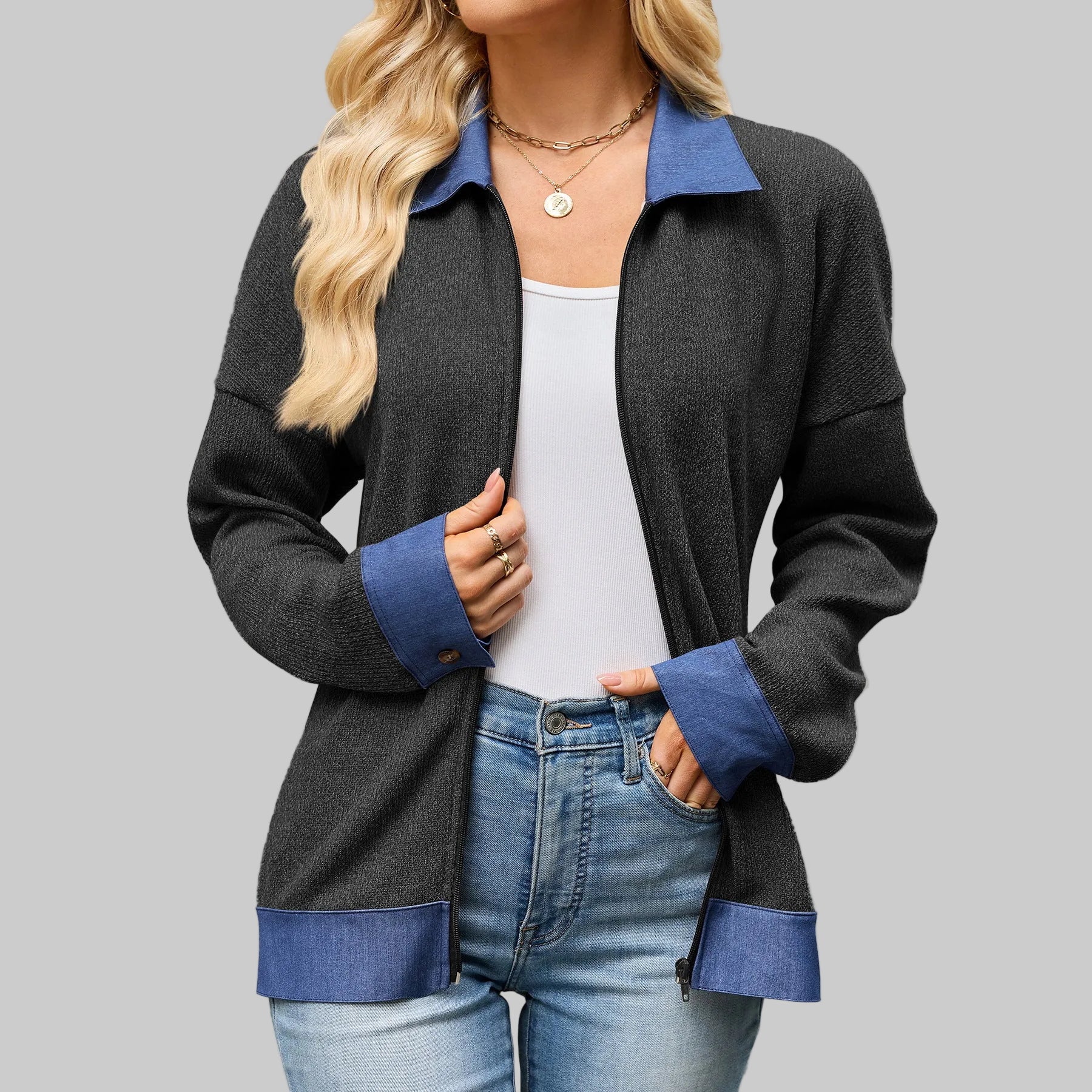 A woman wearing a dark charcoal gray zip-up cardigan with contrasting blue collar, cuffs, and hem, styled over a white tank top and blue jeans.