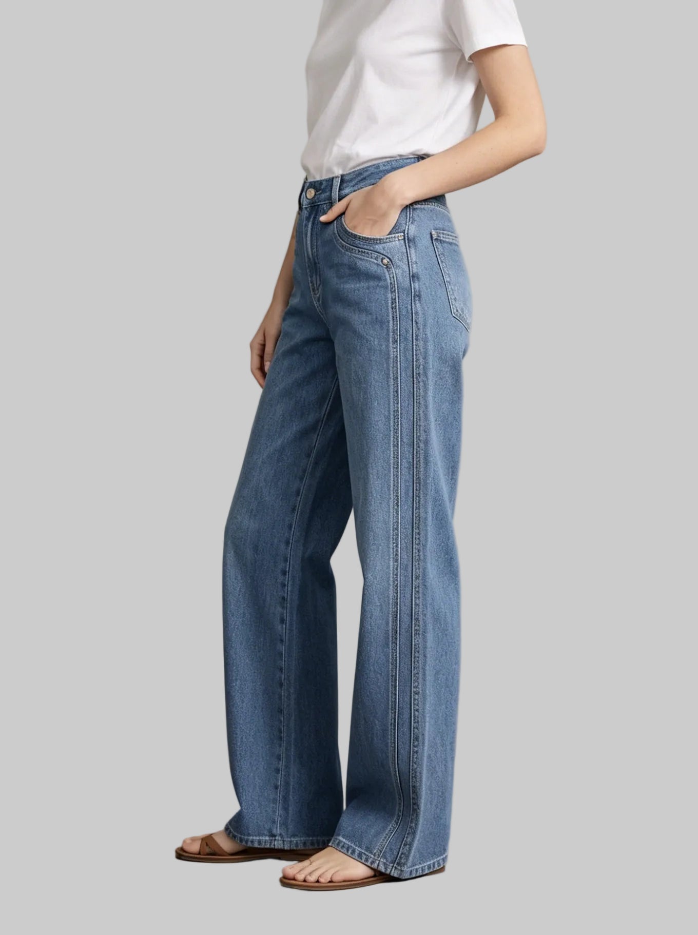 A model wearing high-waisted, wide-leg blue denim jeans with distinctive vertical contrast stitching down the outer legs, styled with a white t-shirt and flat sandals against a textured gray studio background.