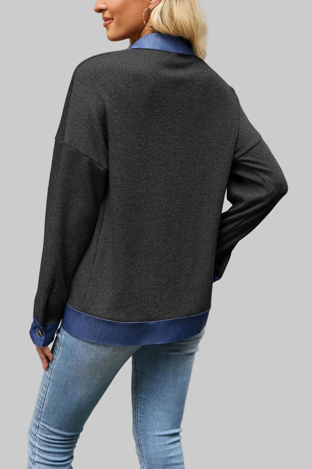 A woman shown from the back wearing a dark charcoal gray knit cardigan with contrasting blue ribbed collar, cuffs, and hem, paired with blue jeans, standing outdoors against a beige stone wall.
