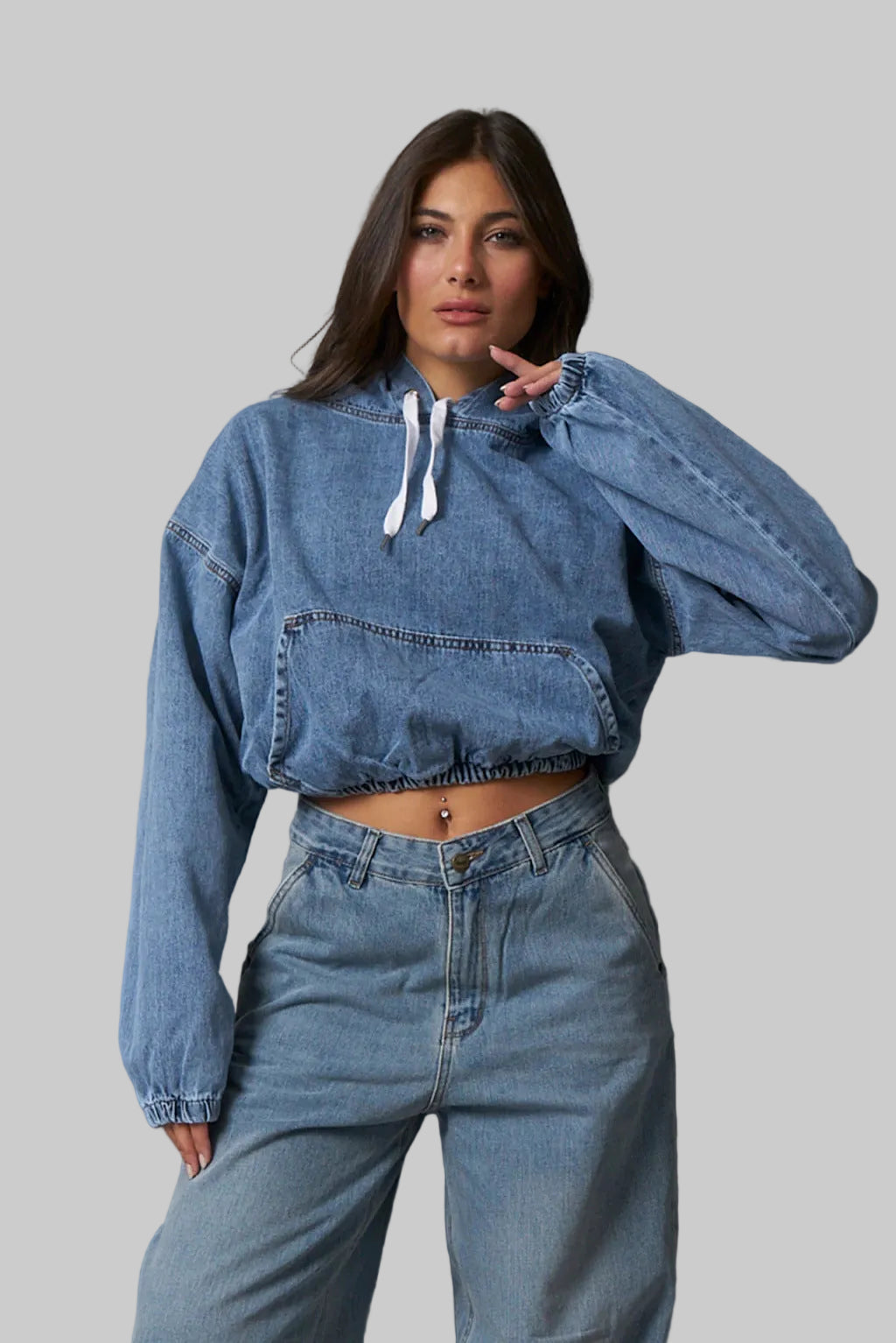 Woman wearing a blue denim hoodie and jeans against a gray tiled wall.