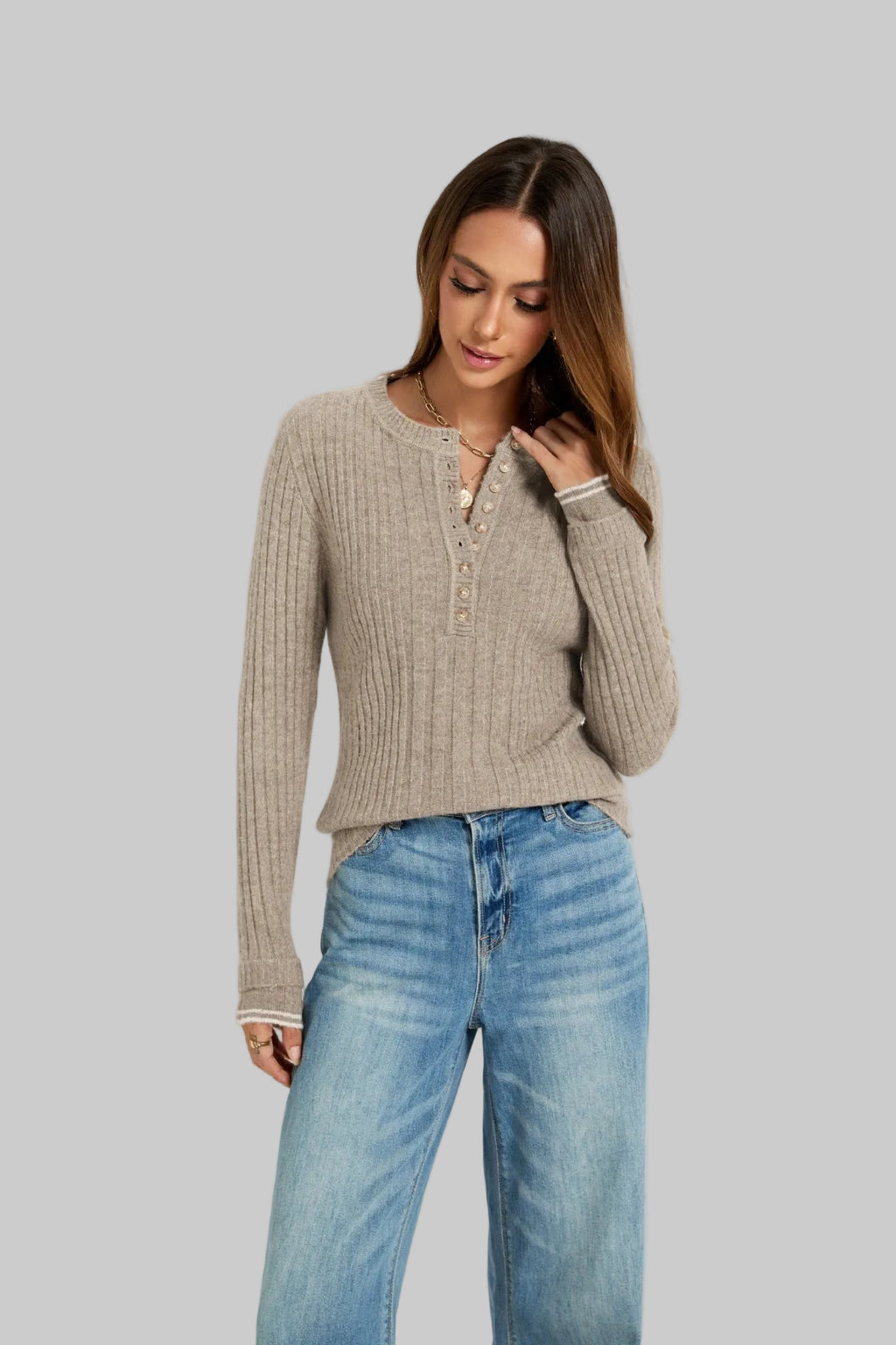 A woman wearing a beige ribbed knit henley sweater and light blue jeans, standing against a plain light gray studio background.