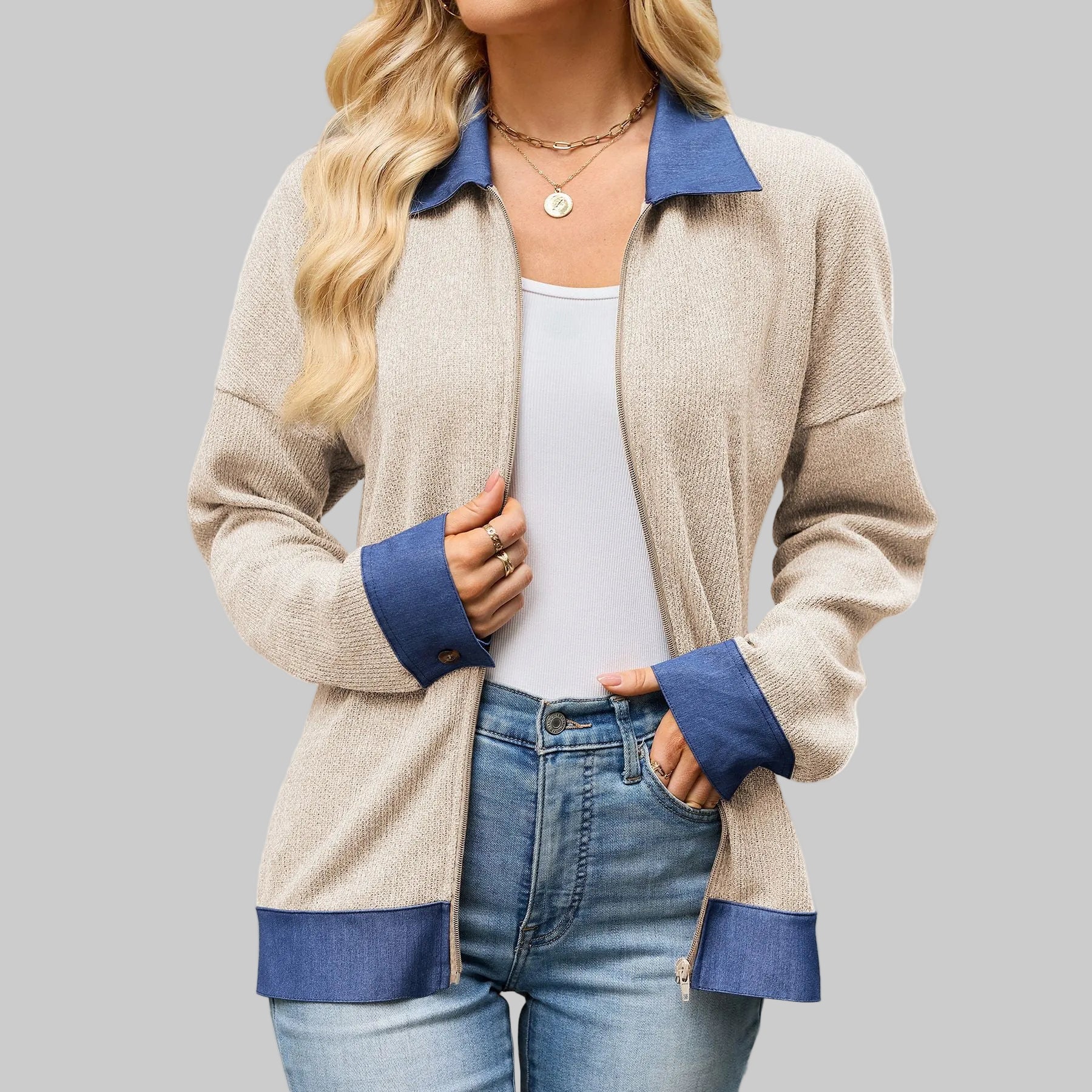 A woman wearing a beige textured zip-up cardigan with contrasting blue collar, cuffs, and hem, paired with a white tank top and blue jeans against a white background.