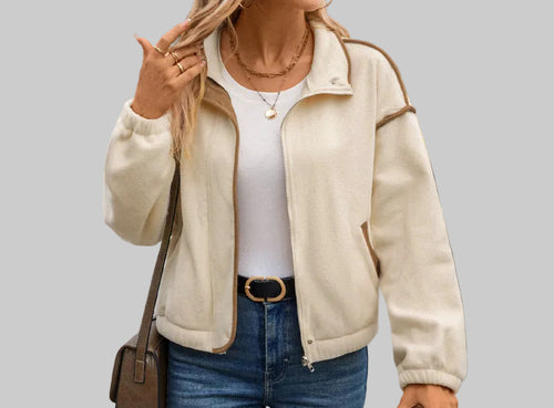A woman wearing a cream-colored zip-up jacket over a white top and blue jeans, carrying a brown shoulder bag and holding a coffee cup in an outdoor setting.