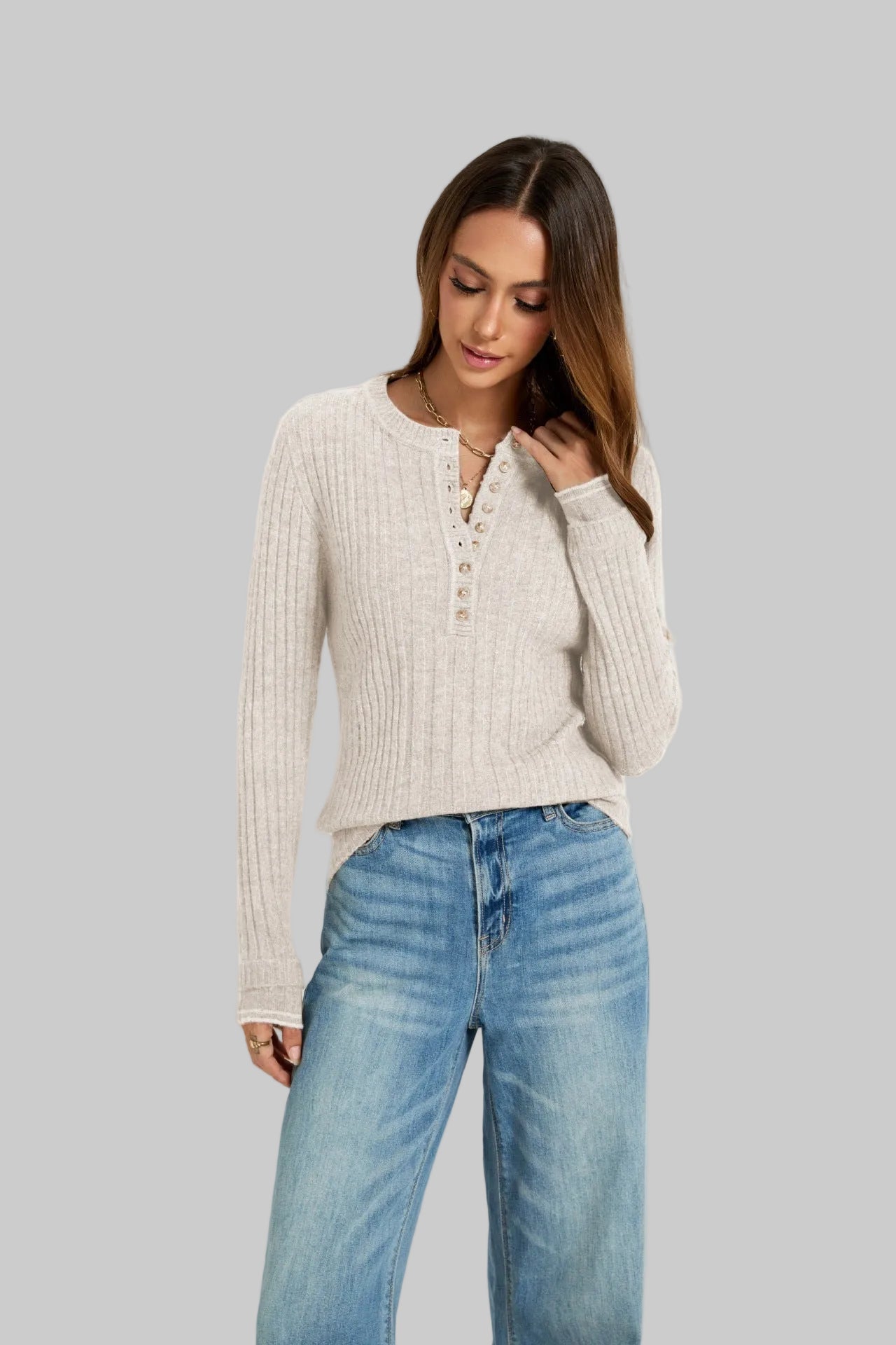 A woman wearing a ribbed knit long-sleeve henley top in a light cream color, paired with light blue jeans, photographed against a plain light gray studio background.