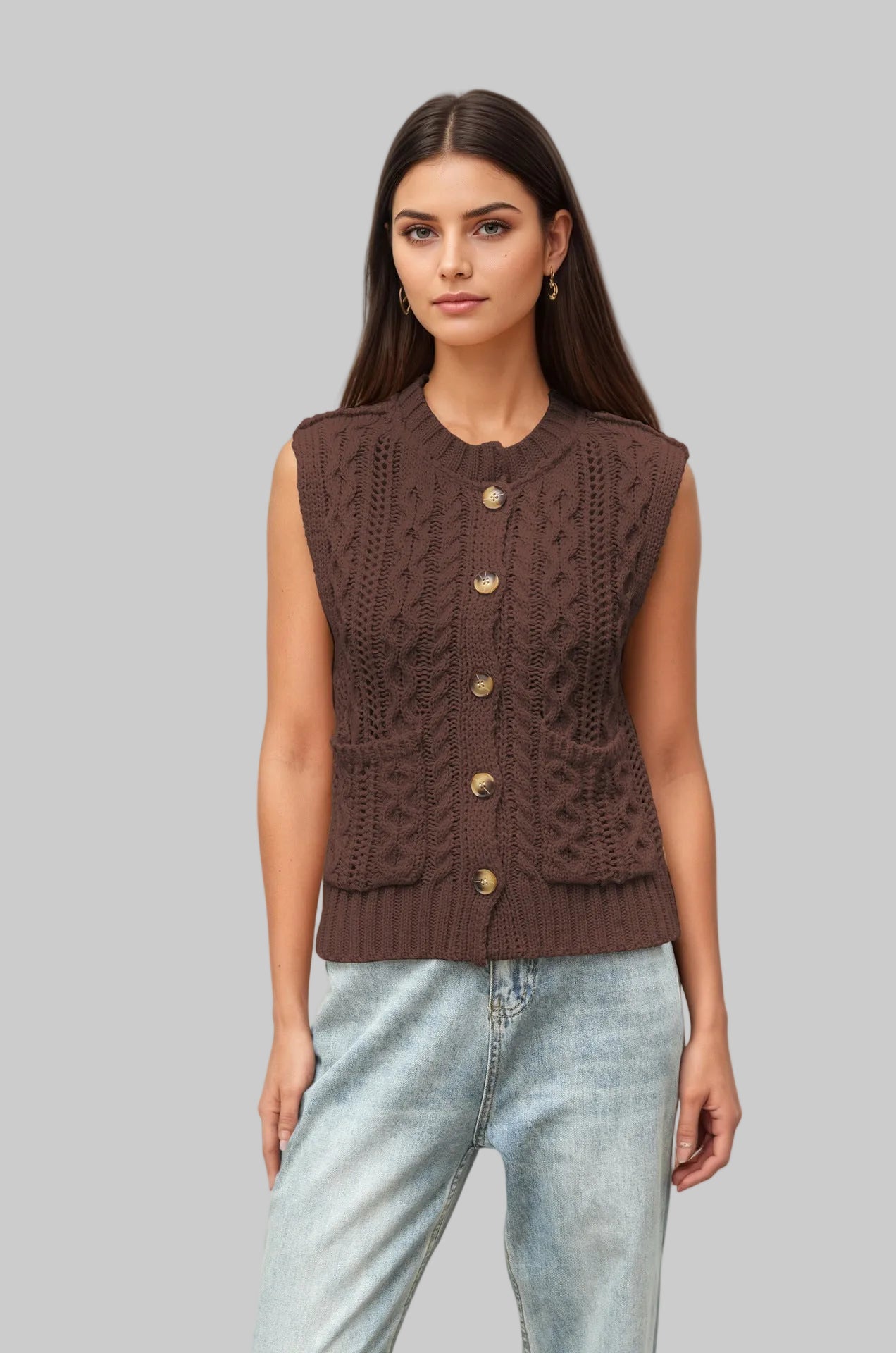 A woman modeling a sleeveless brown cable-knit vest with gold-tone buttons, paired with light-wash blue jeans, against a solid light beige background.