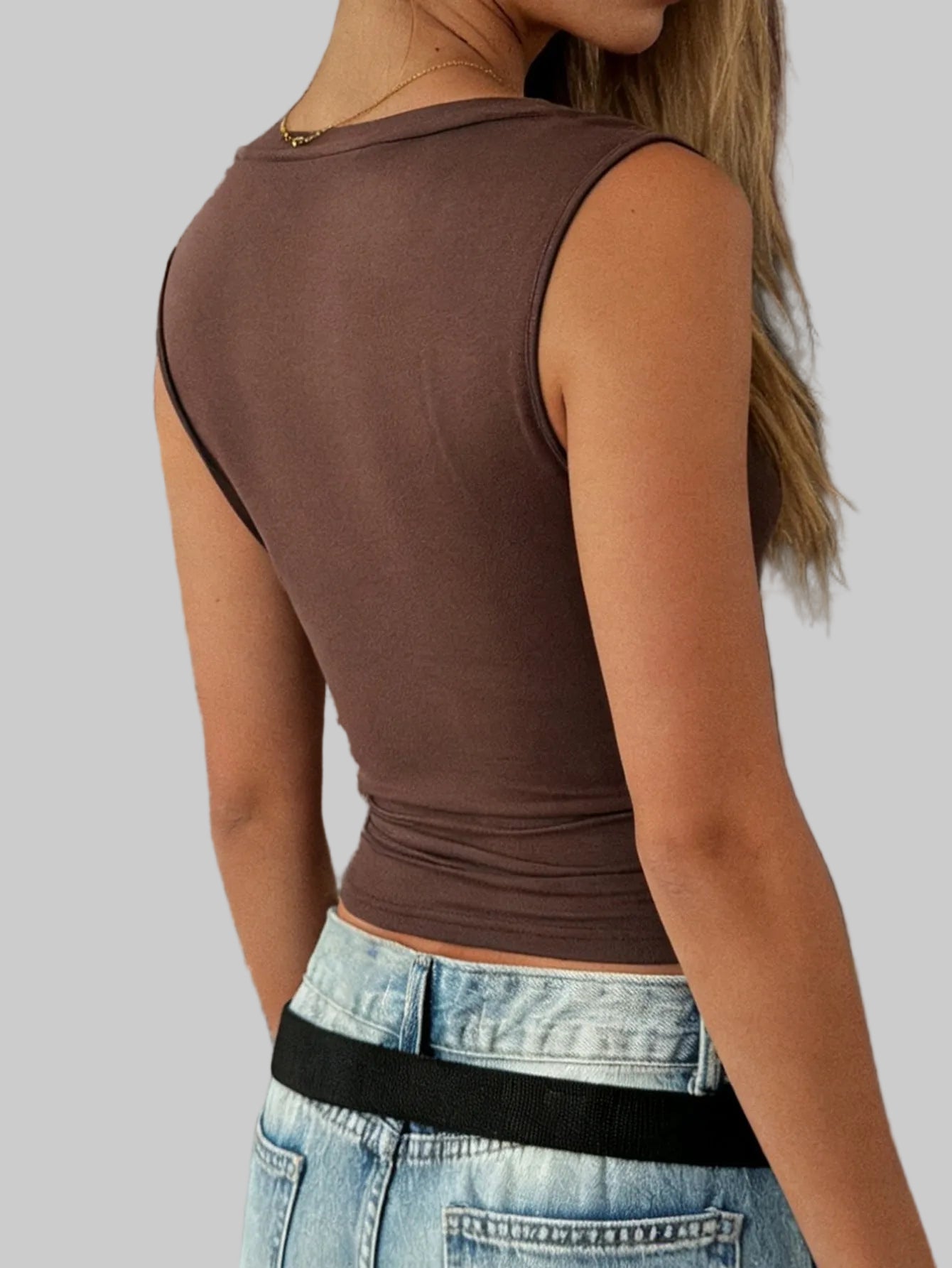 Person wearing a brown sleeveless top and blue jeans against a stone wall.