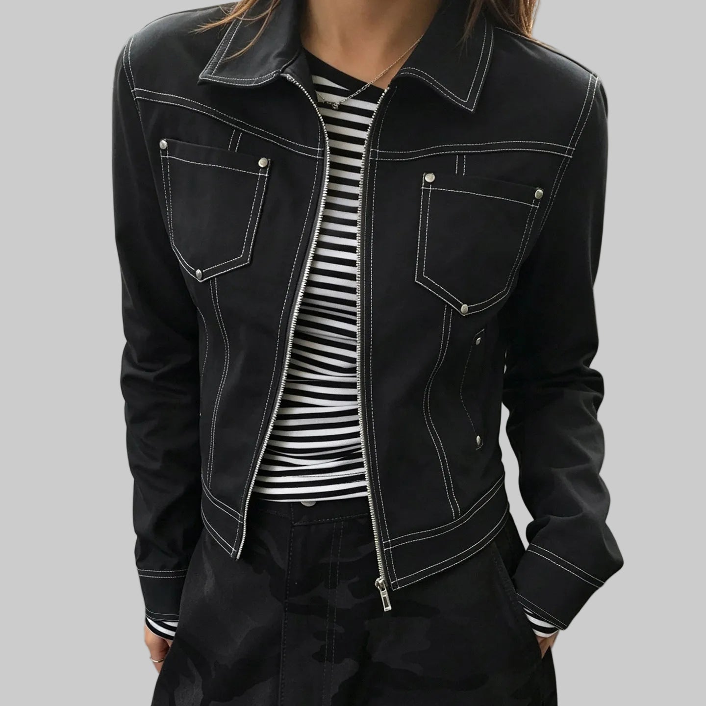A person wearing a black cropped denim jacket with white contrast stitching, silver snap details, and a front zipper, layered over a black and white striped shirt.