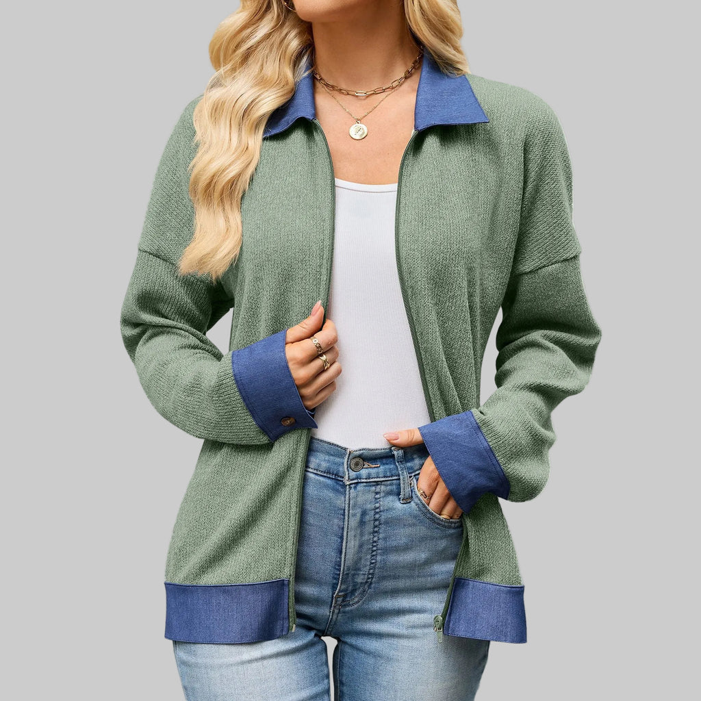 A woman wearing a sage green knit zip-up cardigan with contrasting blue collar, cuffs, and hem, styled over a white tank top and blue jeans against a plain white background.