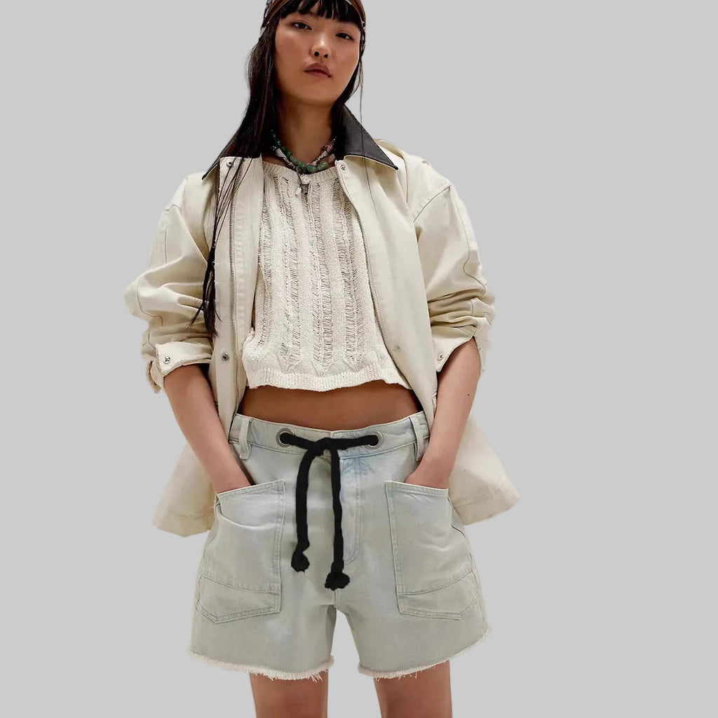 Woman wearing a beige jacket, white lace top, and light blue shorts on a plain background
