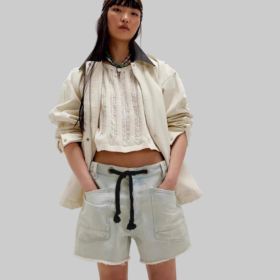 Woman wearing a beige jacket, white lace top, and light blue shorts on a plain background