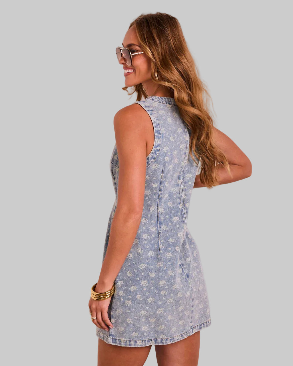 Woman wearing a light blue floral dress holding pink flowers against a white wall.